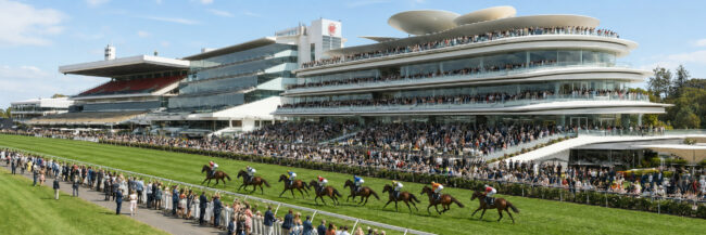 Wide view of a horse race at a modern grandstand filled with spectators on multiple terraces along the track.