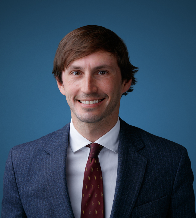 Professional headshot of a man in a navy pinstripe suit and burgundy tie, smiling against a blue studio backdrop.